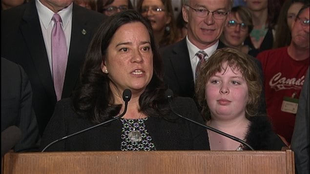 Justice Minister Jody Wilson-Raybould stands at the podium in the Foyer of the House of Commons Tuesday to announce federal legislation that would guarantee legal and human rights protection to transgender people in Canada.  Charlie Lowthian-Rickert, 10, born a boy but who identifies as a girl, joined the minister for the announcement. The raven-haired justice minister stands at the podium speaking into two mics. She wears a dark sweater and her chin is thrust forward in a look of determination. To her left is a young woman with blond, curly hair and a slightly ruddy complexion, who is listening attentively.