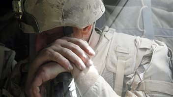 Many Canadian veterans are still waiting for jobs they were promised. We see a closeup of the face of a soldier dressed in a camouflage battle helmet. His gaze is downward and he wears a sad expression. His left hand is crossed over his right with his palms.