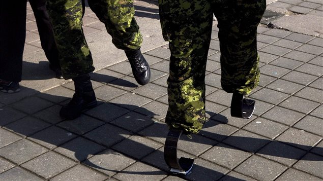 Master Cpl. Jody Mitic walks, on his 'running legs' past a fellow soldier, as he prepares for a charity run in Toronto in 2009. Many veterans are chagrinned about the slow pace of hiring injured colleagues. We see the legs of two soldiers who are wearing camouflage pants. The soldier to the left wears regular combat boots. The runner on the right wears springy blades (like those worn by former South African runner Oscar Pistorius) at the bottom of his pants.