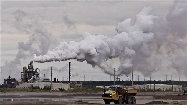 A dump truck works near the Syncrude oil sands extraction facility near the city of Fort McMurray, Alta. in 2014. A new study in the journal Nature finds that Alberta's oil sands are one of the largest sources of organic aerosol air pollution in North America.  We see an enormous yellow dump truck in the foreground of the photo. Behind it stands a grey factor from whose smokestack white and grey smoke is billowing up toward a dark sky. Think of the moon having a bad day.