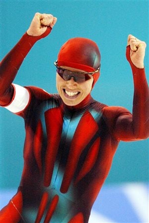 Le May Doan after winning the gold medal in the women's 500m Olympic speed skating competition at the 2002 Salt Lake City Olympics. She has both hands raised in the air in triumph and a wide, glowing smile on her face. She is dressed in the red and black racing suit and a black helmet.