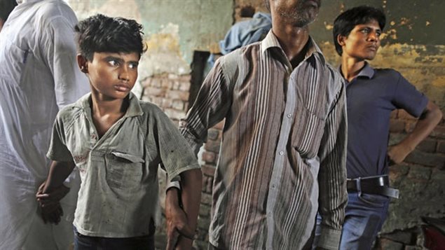 Workers from the Save the Childhood Movement remove a bonded child labourer after a raid at a factory in New Delhi, India in 2012. We see a boy (perhaps 11 years-old) to the left of the photo. He has black, tousled hair and fiery eyes and wears a drab grey shirt. To his left are two adults, one in a grey, stripped shirt and the other in a blue polo shirt. The man in the stripped shirt is holding the boy's left arm and appears to be leading him.