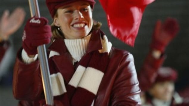 Catriona Le May Doan carried the Canadian flag at the 2002 Olympic Winter Games in Salt Lake City. We see her dressed in her red jacket and red and white scarf looking to the left of the photo. She holds the staff of the staff of the flag in her right hand and it rises over her right shoulder. She has something of forced smile on her face.