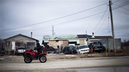  man rides his ATV in the northern Ontario First Nations reserve in Attawapiskat, in April after the community's chief declared a state of emergency because of suicide attempts. We see the man dressed in a black snowsuit atop a red ATV. He rides on a dirt road in front of pair of dilapidated buildings that have five cars and SUVs, two of them in disrepair, in front of them.