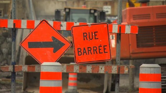 Canadians fight to save their sanity with--seemingly--every road they turn to. We see orange band black signs above an orange and white barrier. The sign on the left holds a big, black arrow pointing to the right. The sign next to it reads in French, "Road Blocked."