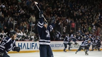  The stands were full in Halifax as forward Holden Cook of St. Francis Xavier University celebrates after his game-winning goal in the third overtime at Canada's university championships this spring. We see a young man in a dark blue hockey uniform with a big white X on the chest. His stick is high in the 
