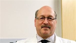 Dr. Mark Freedman, a neurologist, is the director of MS research at The Ottawa Hospital and a professor at the University of Ottawa. We see a large, balding man wearing glasses from the bottom of his chest up. He wears a dark tie under his white lab coat. He projects the air of a friendly person. 