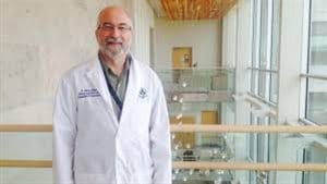 Dr. Harold Atkins is a stem cell transplant physician and scientist at The Ottawa Hospital and an associate professor at the University of Ottawa. We see a man standing with the top of an atrium behind him. He wears glasses and a lab coat and is smiling a friendly smile. He is greying and beginning to lose his hair.