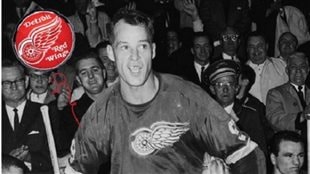 Gordie Howe in his prime in the 1950s. Howe spent most of his NHL career with the Detroit Red Wings, and played 33 pro seasons. It's a black and white photo with Howe, missing most of his teeth, in a dark Red Wings jersey. He is looking up the ice under hair that his beginning to recede. We see his famous number nine on the sleeves of the jersey. Behind him we see the fans dressed in dark suits and ties and--in 1950s fashion--wearing short hair.