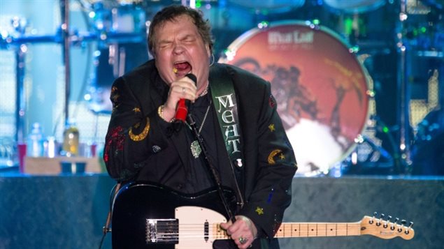 Meat Loaf, seen in a 2013 European show, is best known for his 1977 album 'Bat Out of Hell."  We see a large man in a black jacket with gold decals or embroidery of moons and stars on it. He is sitting and singing into a microphone, holding the mic with his right hand. His hair is short and his mouth is wide open. His face appears flushed. Behind him we see a large drum kit but not the drummer.