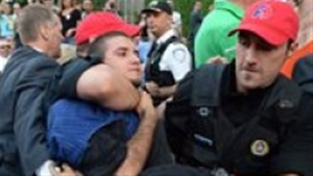 Transgender activist Esteban Torres was quickly detained by police after an altercation with Quebec Premier Philippe Couillard Thursday in Montreal. We see a man who appears to be in his 20s wrapped in the arms of two policeman wearing red baseball caps and dark blue uniforms. The officer to the left has his large forearm around the man's neck. The other officer is holding the man's left arm. We see another officer and part of the crowd in the background.