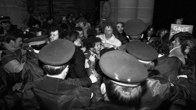 A man scuffles with police outside the Ontario Legislature on Feb. 6, 1981, after about 1,000 gay rights demonstrators marched there in protest of the bathhouse raids. On Wednesday, Toronto's current police chief apologized for the raids. It's a black-and-white photo. We see several men surrounded by police. One man in the middle is bleeding across his face. We don't much see the faces of the police. Rather we see the back of their military hats. The atmosphere appears pitched and extremely tense.