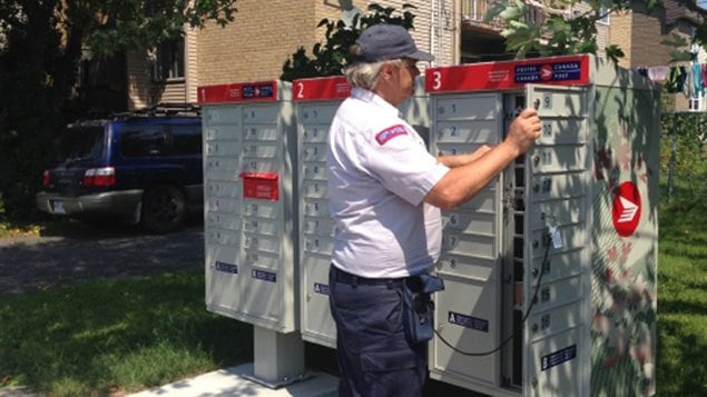 Negotiations to keep postal workers on the job continue after Canada Post  backtracked on its threat to lock out its workers on Monday and said it hoped the Canadian Union of Postal Workers would not issue a strike notice. We see a middle-aged, white-haired postal worker wearing a blue baseball cap and a Canada Post ensignia on the right sleeve of his white shirt (atop blue pants). He has the right end of one of those community postal boxes wide open as he prepares to put letters (and the like) in the individual mail boxes inside the larger box. We see the key to the box at the end of a cord that extends from the postman's belt to the beige box. At the right end of the box, we see the red and white symbol of Canada Post.