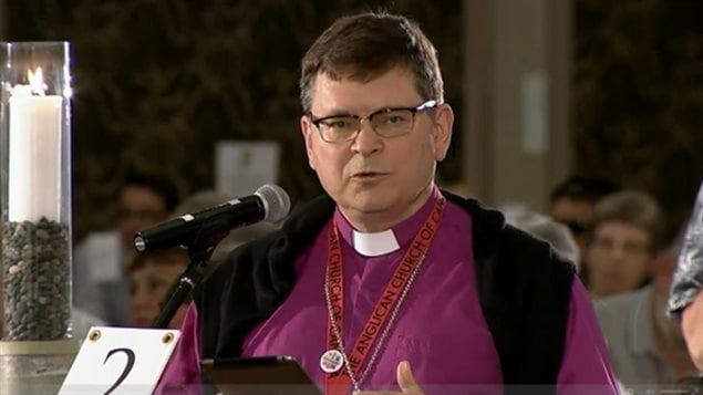 Toronto Archbishop Colin Johnson plans to perform same-sex marriage in defiance of a Monday vote by the Anglican hierarchy. The bishop is a stocky man with short, dark hair combed from a part on the left. He wears glasses that are dark at the top and rimless at the bottom. He stands a microphone wearing a white clerical collar over a red shirt and black sweater. Down his chest are the two straps (a lighter red than the shirt) holding what is likely a plastic pass/i.d.