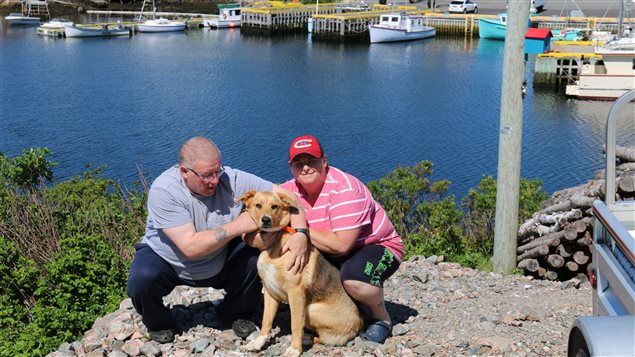 Normn Strickland, his daughter Bet, and their dog, Sadie, are still pondering their confrontation with a pod of killer whales. We see Strickland on the left and Bet on the right. They are croached on a rock with Sadie who appears mostly a Golden Lab. Both are powerful figures and are dressed in summer casual shirts. Bet wears a red Montreal Canadiens baseball hat. Behind them is blue water with small boats sitting at docks. 