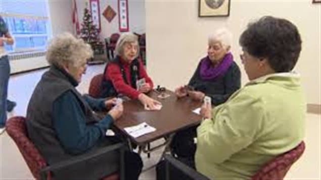 A new survey suggests that retired Canadians are among the happiest in the world--ranking second only to retirees in Mexico. We see four elderly ladies sitting at the four sides of a card table. It appears to be an old-age home as they is a wide window behind them with a tiny Christmas tree on a shelf. The ladies are dressed warmly in sweaters as the one in the centre sets down a trump.