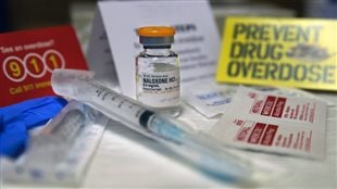  Firefighters in Vancouver and Surrey now carry overdose-reversing naloxone in their bid to cut down the increasing number of drug-related deaths, many of them linked to fentanyl. We see a syringe lying on a table with a bottle marked naloxone behind it. At the edge of the table are two signs. One in yellow says 