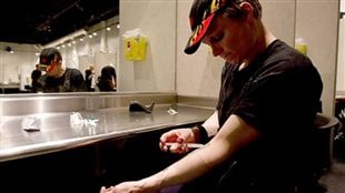 Authorities are doing the best to get users to shoot up only at safe injection sites to avoid drugs mixed with fentanyl. We see a male of about 30 dressed in a baseball cap and dark shirt. He is injecting drugs into his left forearm just above the elbow. He is sitting at something resembling a dressing room table. There is a long shelf with a mirror at its edge.