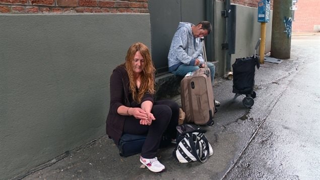 'Exile' says she overdosed on what she believes was fentanyl without knowing it was in the drugs she was taking. We see a red-haired female who could be 20 or could be 40. She is dressed in black except for white sneakers. She is sitting with her back up against an outside greenish wall and appears to be switching pills from one hand to another. To her left, a dark-haired man with short hair and sideburns appears to sticking something into his arm.