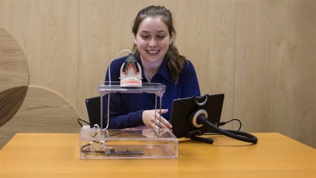 Emma Mogus is pictured with her creation, the Tongue-Interface-Communication (TiC), a tongue controlled computer mouse, in Toronto last Wednesday. We see a lovely girl in a blue sweater with long brown hair sitting behind her invention at a desk.