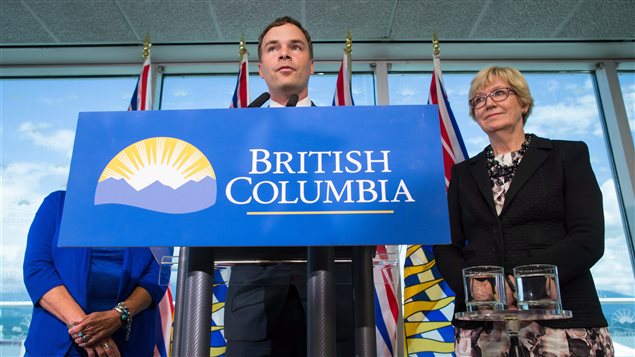 NDP MLA Spencer Chandra Herbert, centre, speaks as British Columbia Minister of Justice Suzanne Anton, right, listens after the government announced it will amend human-rights legislation to protect transgender people, during a news conference in Vancouver on Wednesday. Herbert stands behind a rectangle blue and white sign that reads "British Columbia." It is attached to the podium behind which he speaks. The sign means we really only see him from the neck up. He appears to be about 40 and has short brown hair. Anton, to his left, is a middle-aged woman with large glasses and short blond hair. She is wearing a dark sports coat over a flowery dress.