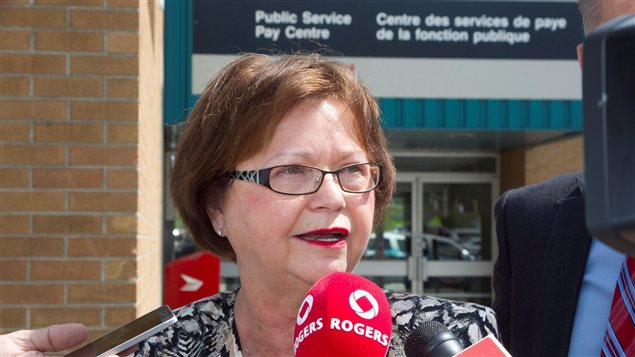 Beleaguered Public Services Minister Judy Foote met the media after touring the government's centralized pay centre in Miramichi, New Brunswick on Wednesday. She's laying some of the blame for problems with the  Phoenix payroll system on a lack of training for staff.  We see a white woman with short brown hair and somewhat fancy glasses speaking into three microphones during a scrum. Behind her is the pay centre, essentially government-issue building brick that identified in English and French.