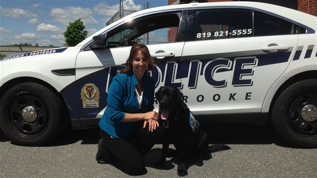  Mélanie Bédard has been with the Sherbrooke police force for 17 years. Kanak joined in May. We see Bédard wearing a blue jacket and black pants on the left. She kneels next to Kanak, who has his mouth open (either from the heat or the fact that he may not like having his picture taken) to the right. Behind them is a white police cruiser with dark blue letters spelling out "Police" and "Sherbrooke."