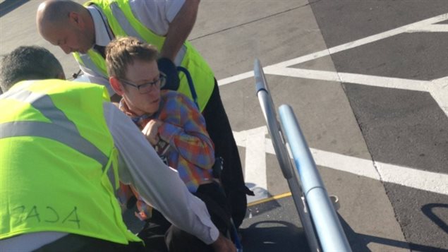Tim Rose is assisted while boarding an Air Canada flight. The airline now says it cannot accommodate his wheelchair. We see Rose in a wheelchair being assisted by two men wearing yellow work vests. He wears glasses and a plaid shirt. His arms are pressed up against his chest.