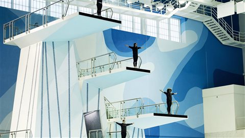 Canadian divers know as the Fab IV, pictured top to bottom, Roseline Filion, Meaghan Benfeito, Jennifer Abel and Pamela Ware pose for a photograph on the diving platform ahead of the winter national diving championships in Scarborough, Ont. in February 2015. They are considered medal contenders in Rio. We see from a distance the four girls dressed in black with their arms extended to the sides. The stand on a series of white platforms, the last of which extends a couple of dozen feet into the air. The walls behind them are coloured various shades of blue.