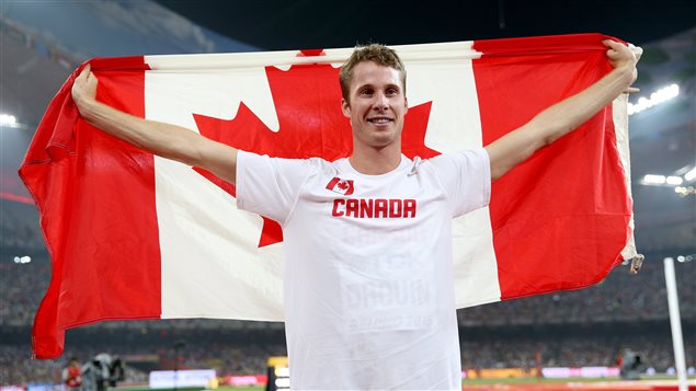 High jumper Derek Drouin won a bronze medal at the 2012 London Olympics  and the gold at the 2015 Athletics World Championships in Beijing last summer (above). He is considered a top contender for gold in Rio. We see a handsome man with short brown hair wearing a white t-shirt with "Canada" and the Canadian flag printed on it. His arms are extended outward like a bird as he holds a large Canadian flag behind him.