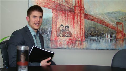 Samuel Fleurent Beauchemin at his office in Victoriaville, QC. We see him dressed in a dark suit at a desk holding a binder. In the background is a large painting of a bridge. The near support of the bridge has a rendering of the photo of the Beauchemin brothers at the top of the story.