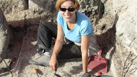 Expedition leader April Nowell in Azraq, Jordan, in 2014. We see an attractive young woman with blond hair wearing sun glasses, a light blue tee-shirt, dark work pants and a wide smile. She holds a trowel in her left hand. A dust pan is to her left as she sits on sand with large, grey rocks behind her.