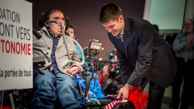 Samuel Fleurent Beauchemin helps Philippe Desrochers, 27, who has Duchenne muscular dystrophy at a Christmas party at which the Fondation le Pont vers l’Autonomie gave out two robotics arms as gifts. We see Beauchemin on the right standing, wearing a dark suit and wide smile below his dark crew-cut hair. He is reaching down with his left hand to adjust something at the foot of Desrochers's wheelchair. Desrochers wears a breathing apparatus and a light sports jacket and grey pants. Attached to the front of the wheelchair is a robotics arm, which is long and thin with what appears to be a tiny computer at its top.