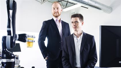 Charles Deguire (left) and Louis-Joseph Caron L'Écuyer, who founded Kinova Robotics 10 years ago, pose beside a JACO robotic arm, designed and constructed at the company's campus. Each wears a dark suit with no tie. Deguire has a light brown beard and his hair is beginning to recede slightly. Caron L'Écuyer is clean-shaven with a crew cut of his darker hair. The robotic arm at the left of the picture is long, dark and shiny. It reaches skyward but another piece--the hand--reaches out from it and is holding a yellow glass.