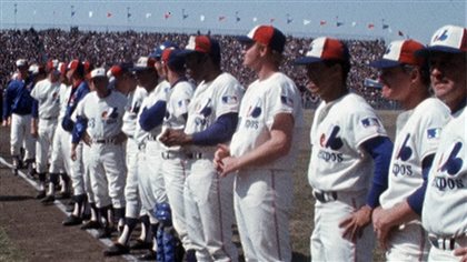 The Montreal Expos line up before their first-ever home game in 1969. We see them in their white uniforms lined up along the first-base foul line with the bleachers of Jarry Park in the background.