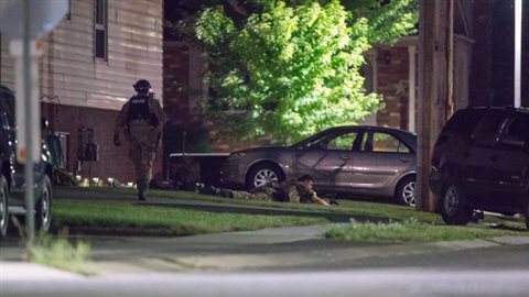 Police kept watch around the Strathroy house on Wednesday night. We see two officers in battle dress--one is lying on the ground, the other approaches a clapboard house. A tree in front of the house is brightly lit. A car and SUV cars sit in front of the house.