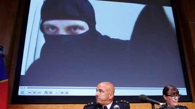 A video of Aaron Driver, who was killed Wednesday in a confrontation with police, is projected on a screen during an RCMP news conference on Thursday. Two RCMP officers (one a bald man, the other a woman wearing glasses) sit at a table. Behind them is a large video of a man with a black robe/scarf wrapped around him and much of his face.