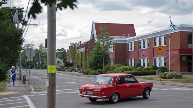 Six male high school students in the quiet community of Bridgewater, N.S. are facing charges following an investigation into complaints that intimate images of at least 20 young female students were shared online without their consent. We see a small red sedan making a turn on an empty street with a woman approaching, walking her bike on a sidewalk. Town looks pretty deserted.