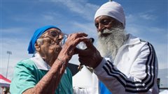 Man Kaur is helped by her 78-year-old son, Gurdev Singh, as she takes a drink of water after competing in the 100-metre track and field event. Man Kaur is helped by her 78-year-old son, Gurdev Singh, as she takes a drink of water after competing in the 100-metre track and field event. They are holding the water bottle together in front of them. Singh is a handsome man who wears a white turban and a long white beard.