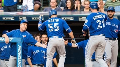 We see the pair (with Martin on the left) about to enter the Jays dugout where they are being greeted by some teammates who are very, very happy. All are wearing blue jerseys and grey pants.