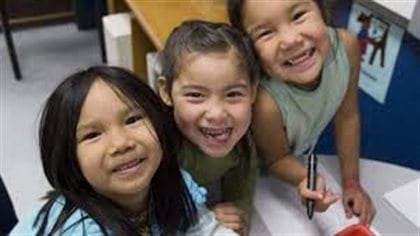 Cindy Blackstock says too many First Nations children are continuing to fall behind children in the rest of Canada. We see three pre-school girls looking up and grinning widely at the camera.