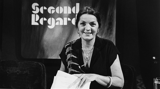 Dans un studio de télévision, l’animatrice Myra Cree, souriante, est assise à un bureau avec des documents dans les mains. Un écran en arrière-plan affiche le nom de l’émission.
