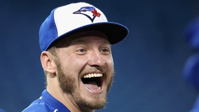 Josh Donaldson, the 2015 AL MVP, is having another superb season in 2016 as the Jays take a serious run to the playoffs. We Donaldson under a white and blue Blue Jays cap. He has long, blondish sideburns (almost 1970s style) and a wide, wide smile across his mouth which is agape.