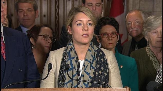 La ministre du Patrimoine canadien, Mélanie Joly, entourée de plusieurs pesonnes pour une annonce devant deux micros.