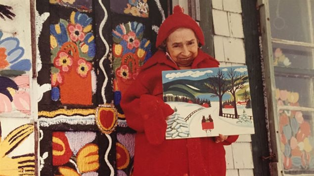 Maud Lewis standing in front of her home with one of her paintings