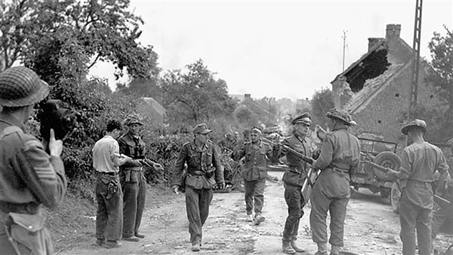 Major David V. Currie (left, wearing binoculars and holding pistol) of The South Alberta Regiment accepting the surrender of German troops at St. Lambert-sur-Dive, France, 19 August 1944. This photo captures the very moment and actions that would lead to Major Currie being awarded the Victoria Cross. Battle Group Commander Major D.V. Currie at left supervises the round up of German prisoners. Reporting to him is trooper R.J. Lowe of *C* Squadron