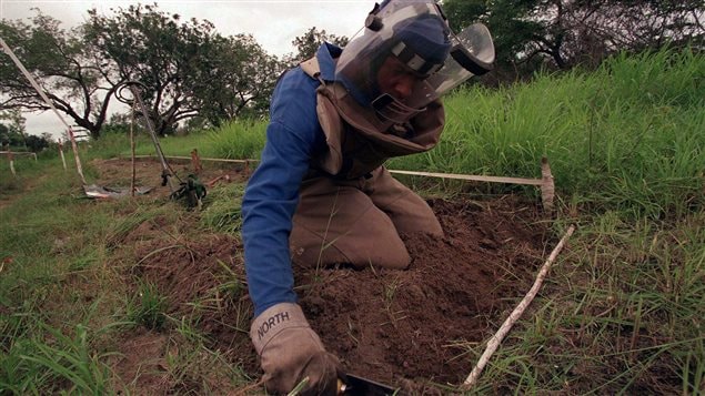 The image shows a demining worker in special gear on his knees probing a ground in front of him for mines.