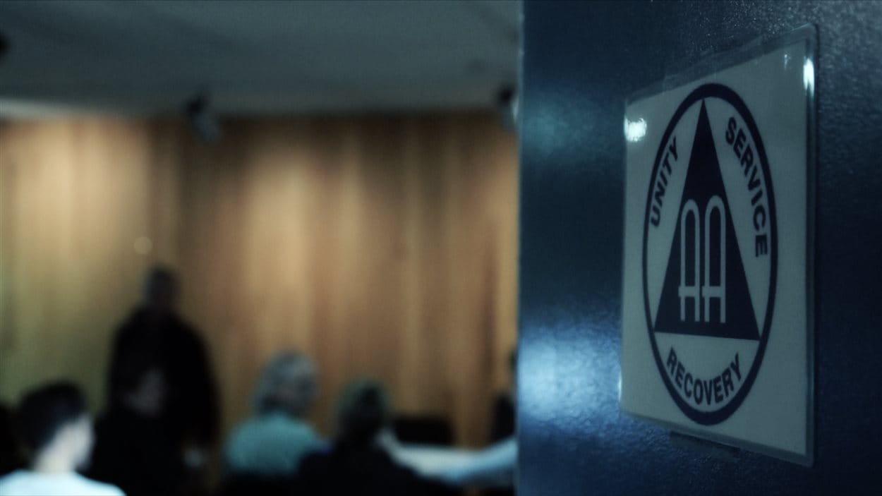 Sur une porte ouverte, un paneau affichant le logo des AA et dans la salle en flou, des gens qui participent à une rencontre.