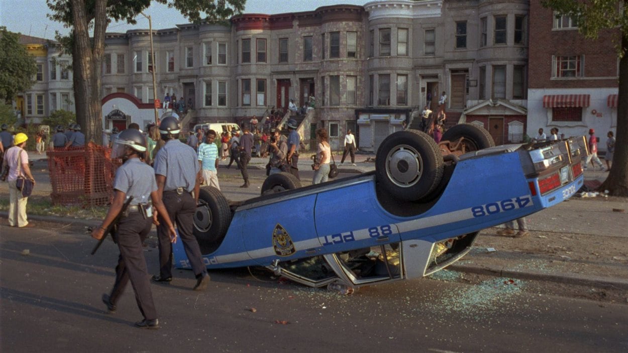 Des policiers marchent près d'une voiture de police renversée dans le secteur de Crown Heights, à Brooklyn, le 21 août 1991, pendant les émeutes qui secouent la ville.