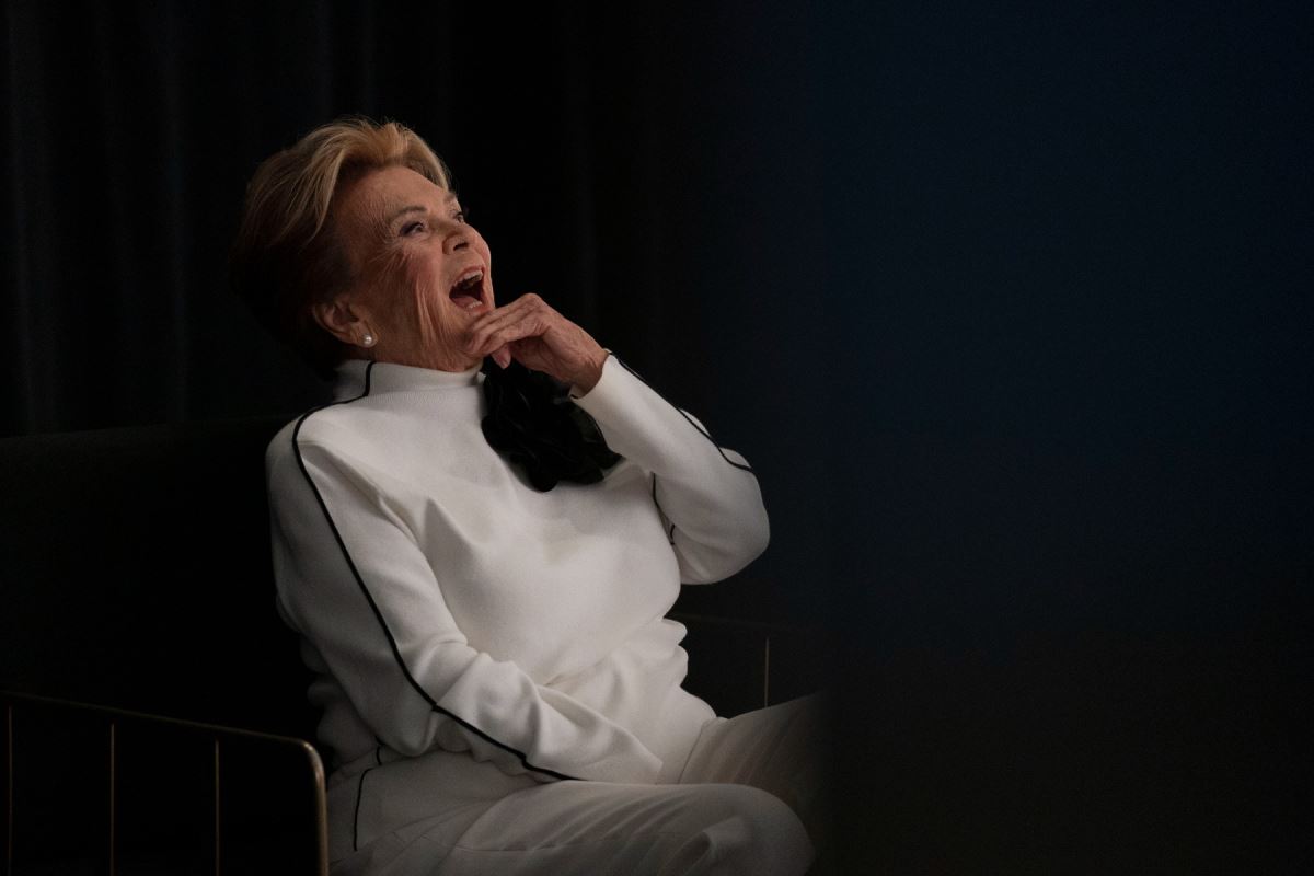 Portrait de Janette Bertrand sur fond noir, riant à pleine dents et se tenant le menton. 